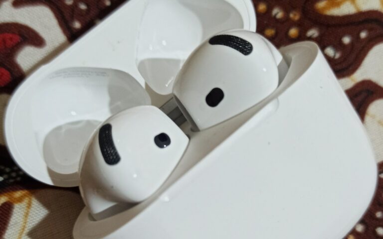 Deux écouteurs Apple AirPods 4 blancs posés dans leur boîtier de charge ouvert, sur un tissu à motifs