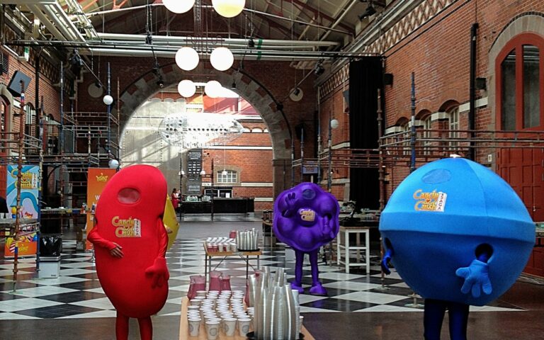 Trois personnes déguisées en bonbons de Candy Crush, rouge, violet et bleu, dans une grande halle en brique