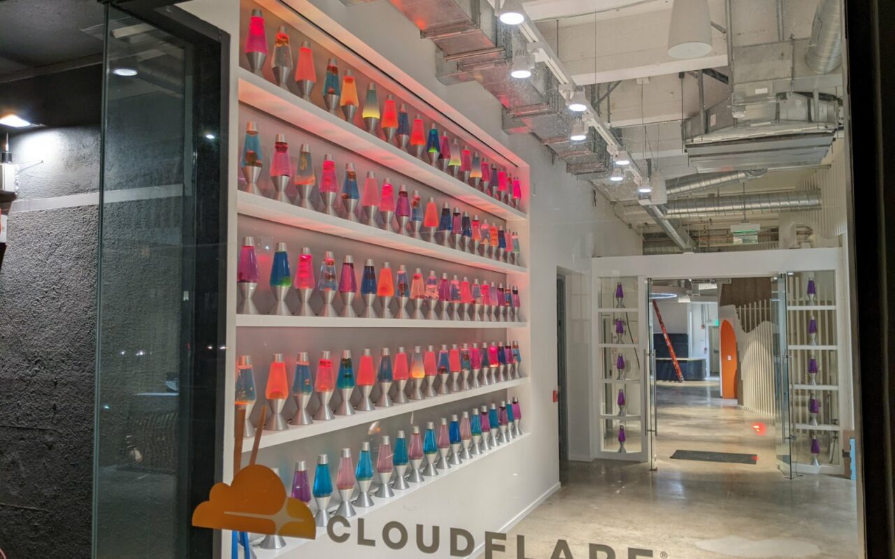Un mur d'étagères blanches garni de dizaines de lampes à lave colorées, avec le logo Cloudflare au pied du mur