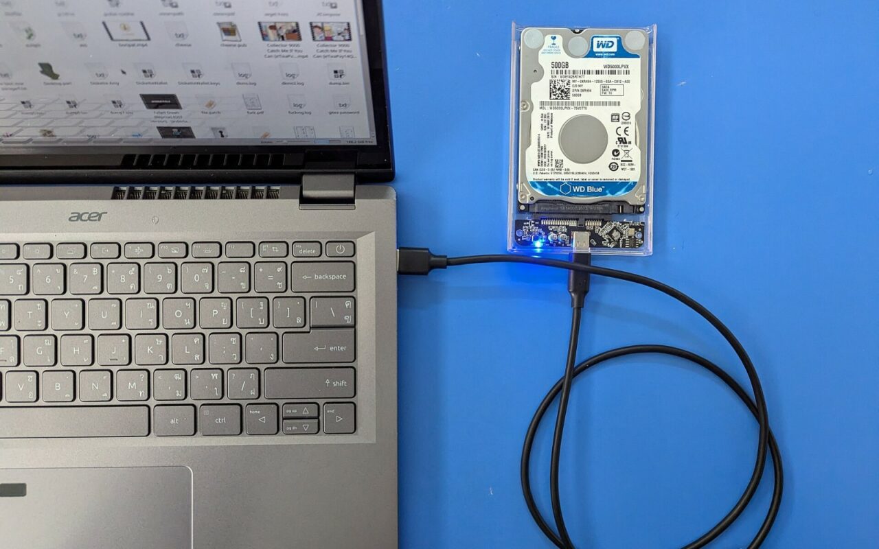 Disque dur 2,5 pouces dans un boîtier USB transparent, relié par un câble à un ordinateur portable posé sur un fond bleu