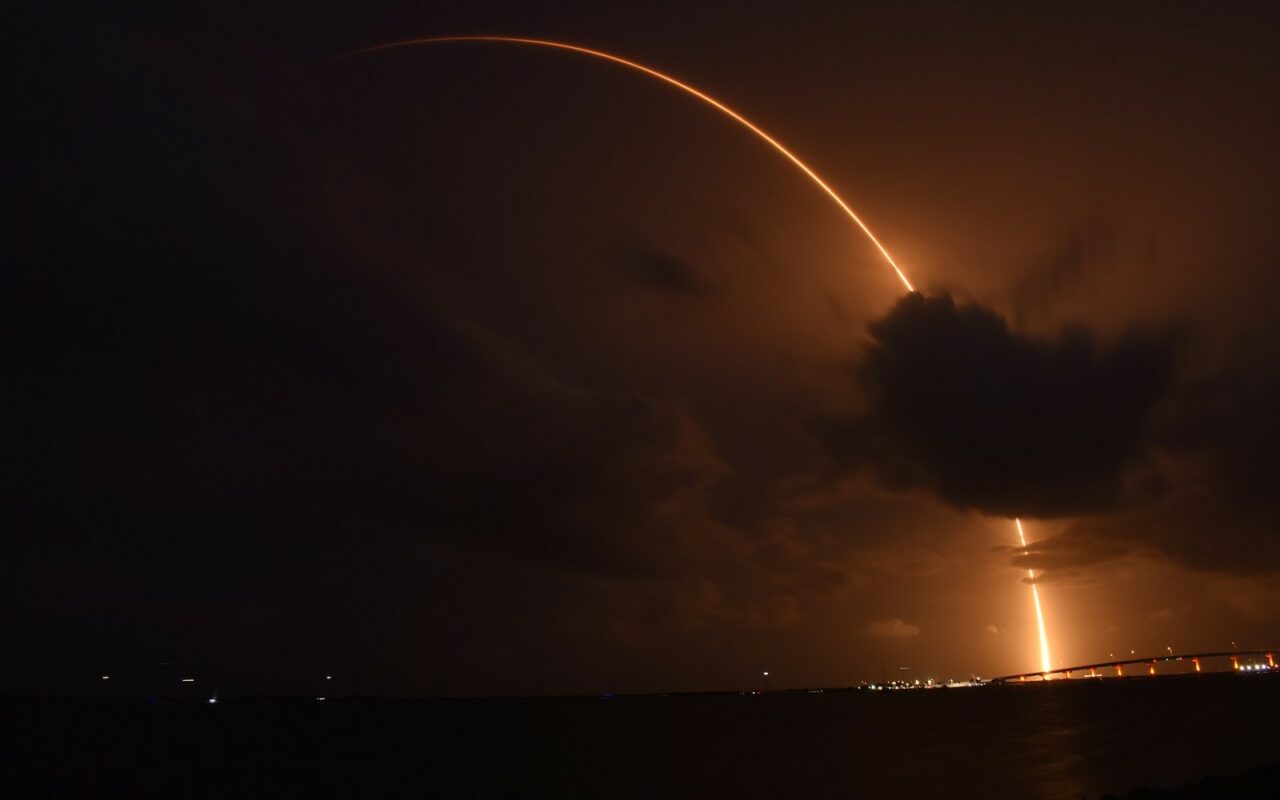 Trainee orange d'un lanceur Falcon 9 en ascension de nuit au-dessus des nuages et du littoral