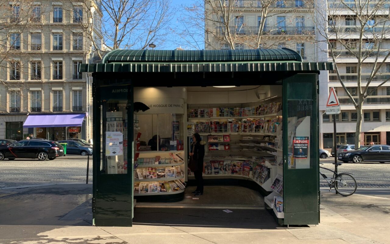 Kiosque à journaux vert ouvert sur un trottoir, présentoirs remplis de magazines et de quotidiens
