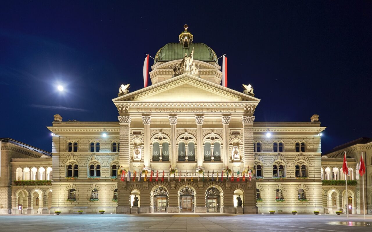 Facade du palais federal suisse eclairee de nuit, drapeaux sur la place