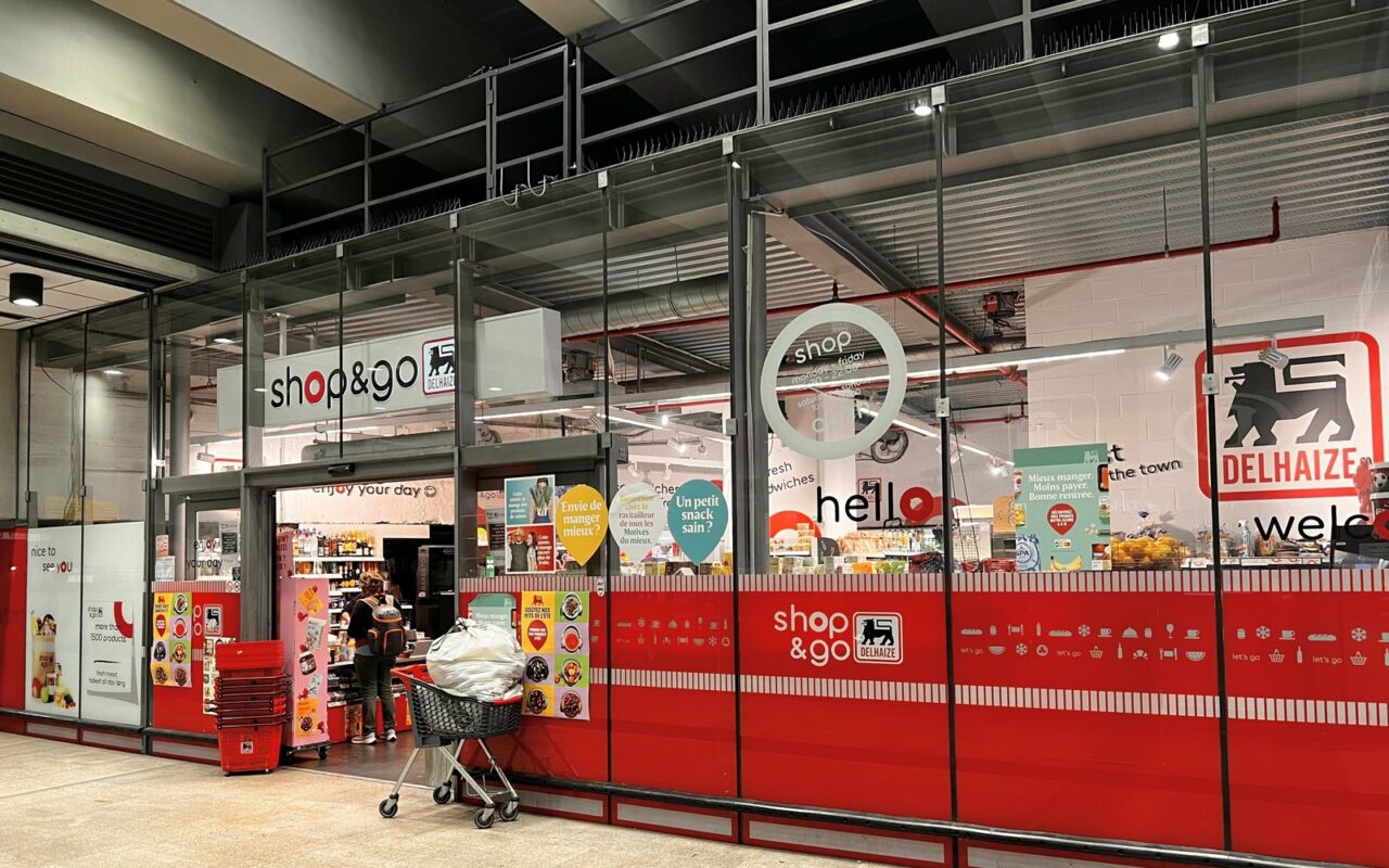Vitrine rouge d'un magasin Delhaize Shop & Go avec le logo au lion, un chariot devant l'entrée
