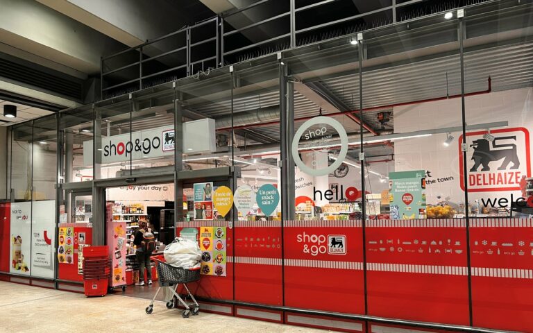 Vitrine rouge d'un magasin Delhaize Shop & Go avec le logo au lion, un chariot devant l'entrée