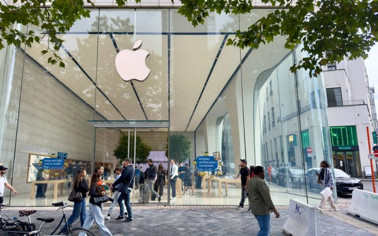 La façade vitrée de l'Apple Store de Bruxelles, surmontée du logo Apple, avec des passants sur le trottoir et des clients à l'intérieur.