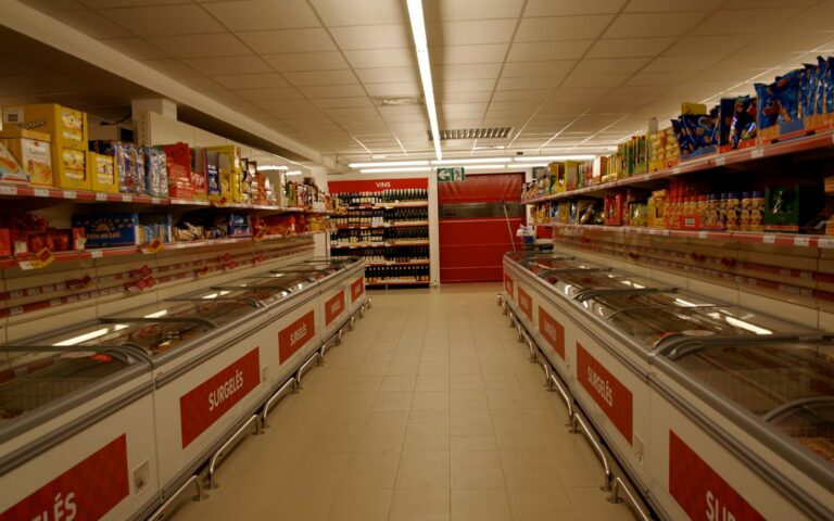 Allée d'un supermarché bordée de bacs de surgelés vitrés, sous des néons, avec des rayonnages d'épicerie au fond.