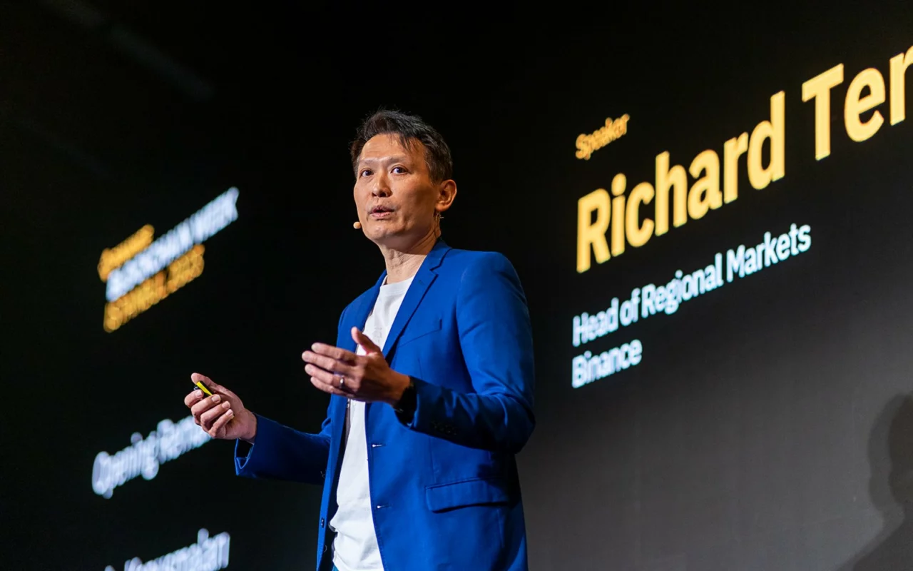 Richard Teng, de Binance, en veste bleue, parle sur une scène sombre devant un écran géant à son nom