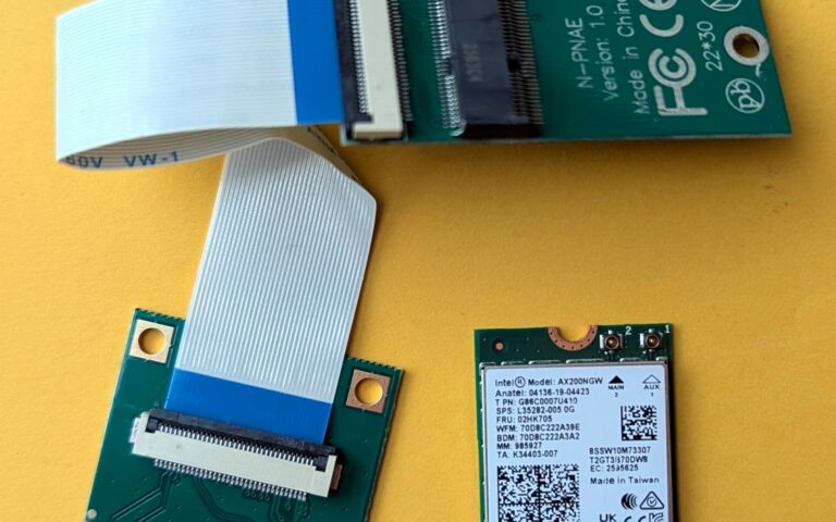 Une petite carte Wi-Fi Intel AX200 au format M.2 posée sur un fond jaune, à côté d'une carte d'adaptation verte et d'une nappe.