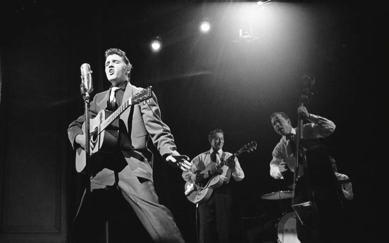 Elvis Presley chante à la guitare sous les projecteurs, accompagné de Scotty Moore à la guitare et de Bill Black à la contrebasse