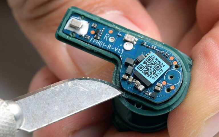 Doigts tenant la carte électronique d'un écouteur Fairbuds 2 ouvert, une lame de cutter glissée sous le circuit