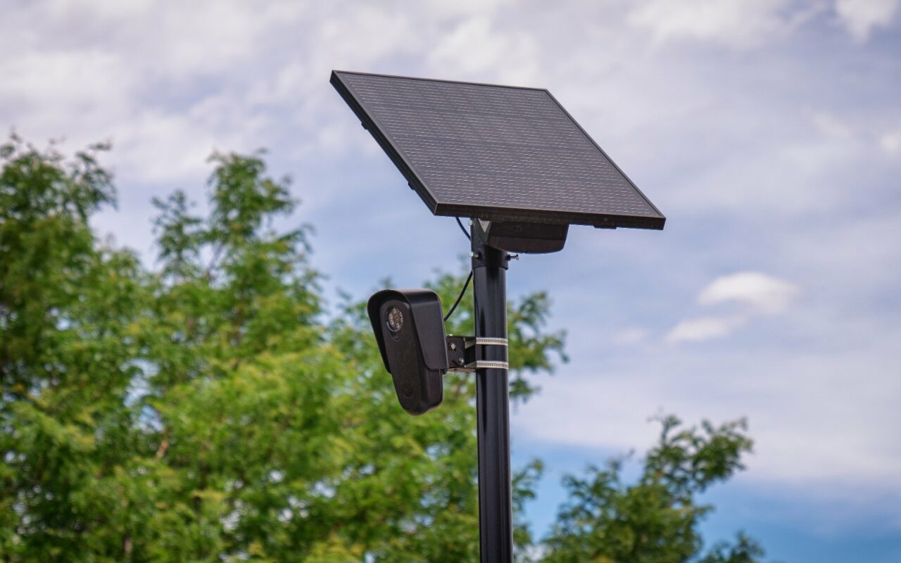 Caméra Flock Safety de lecture automatique de plaques fixée sur un mât noir, sous un panneau solaire, devant des arbres.