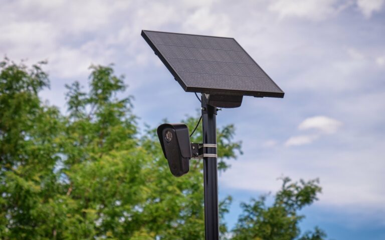 Caméra Flock Safety de lecture automatique de plaques fixée sur un mât noir, sous un panneau solaire, devant des arbres.