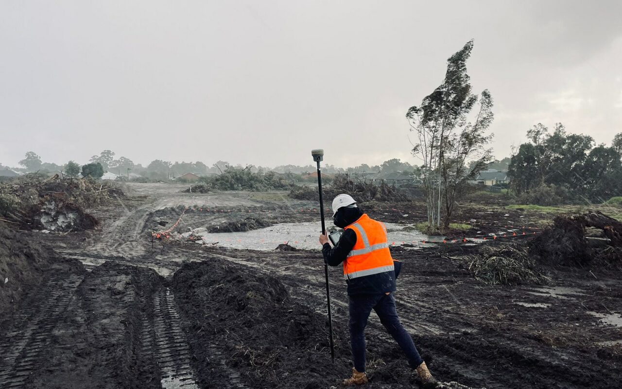 Un géomètre en gilet orange marche sur un terrain boueux en tenant une canne surmontée d'un récepteur de positionnement par satellite.