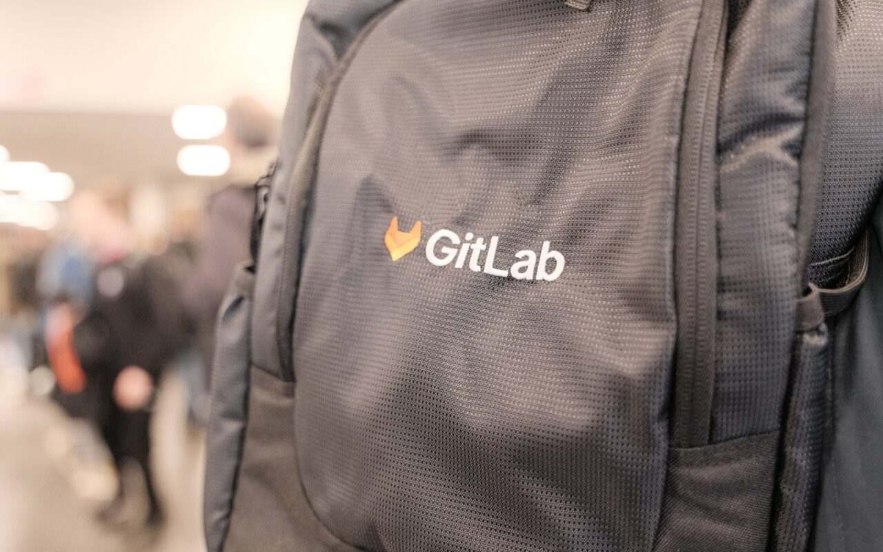 Gros plan sur un sac à dos noir portant le logo orange et le nom GitLab, avec des visiteurs flous dans un couloir du FOSDEM en arrière-plan