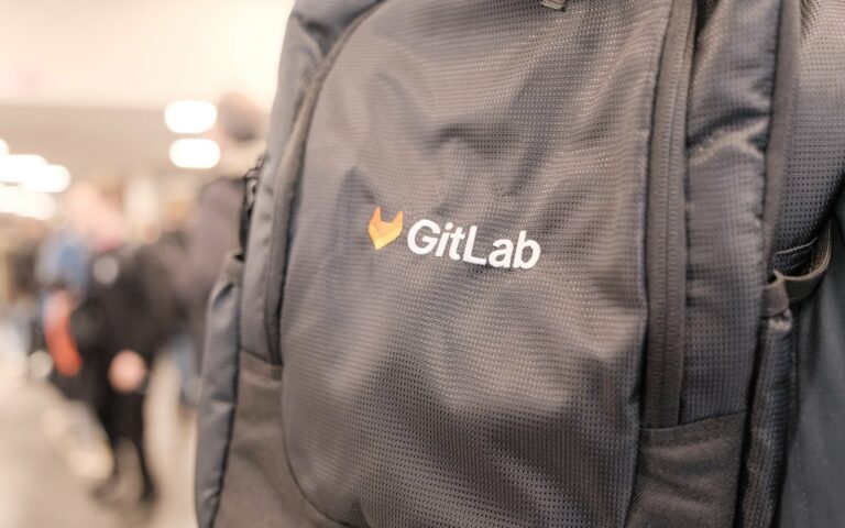 Gros plan sur un sac à dos noir portant le logo orange et le nom GitLab, avec des visiteurs flous dans un couloir du FOSDEM en arrière-plan