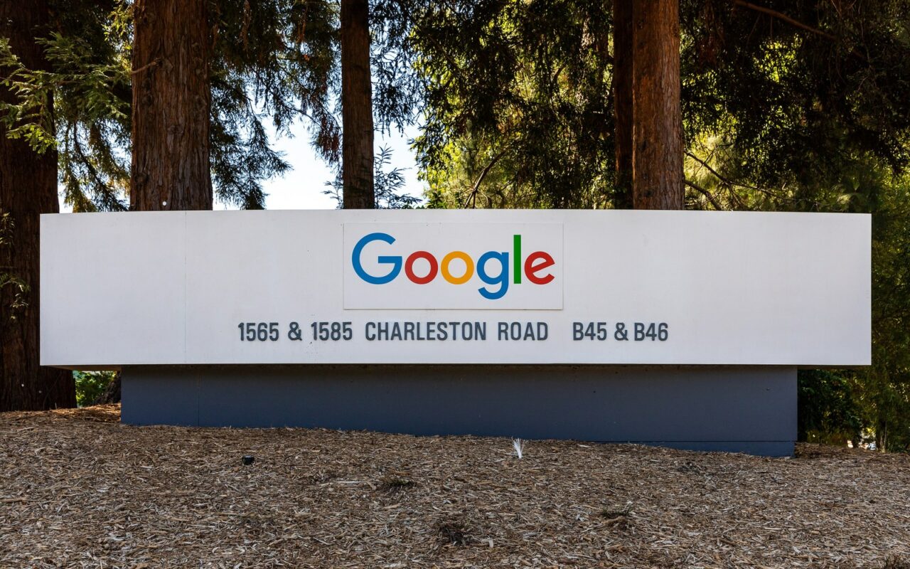 Panneau blanc portant le logo Google devant des bùtiments du siÚge de l'entreprise, entouré de grands arbres