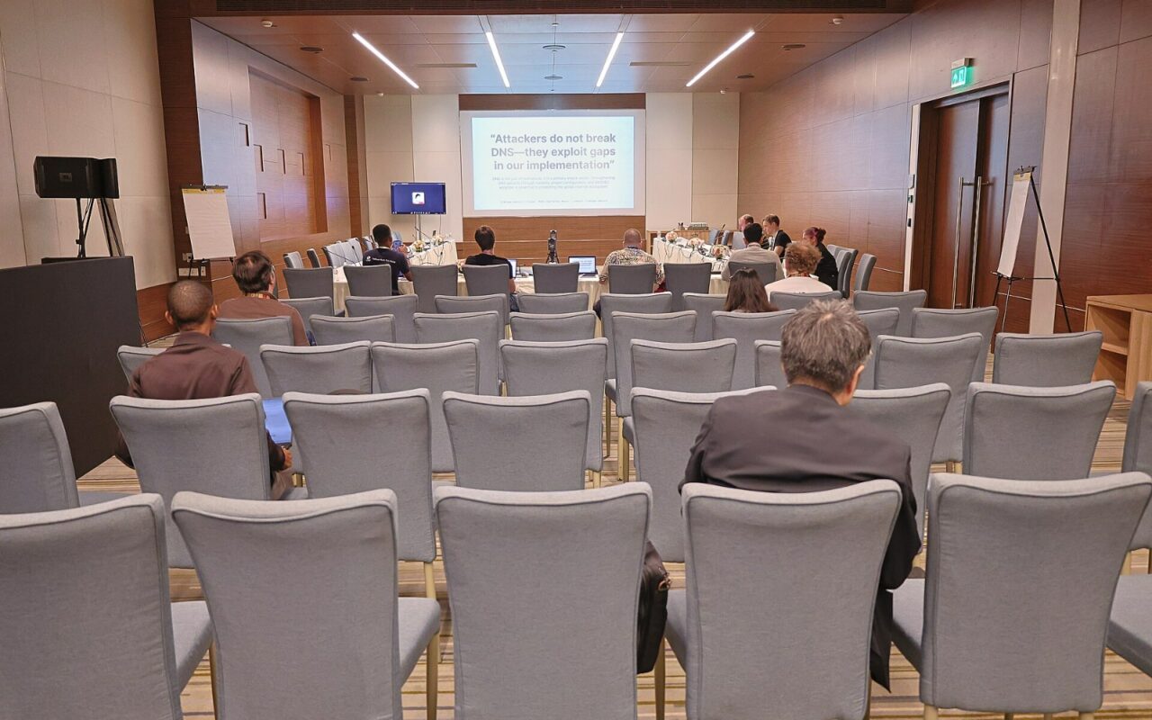 Salle de conférence vue du fond, rangées de fauteuils gris en partie occupées, orateurs à une table sous un écran de projection