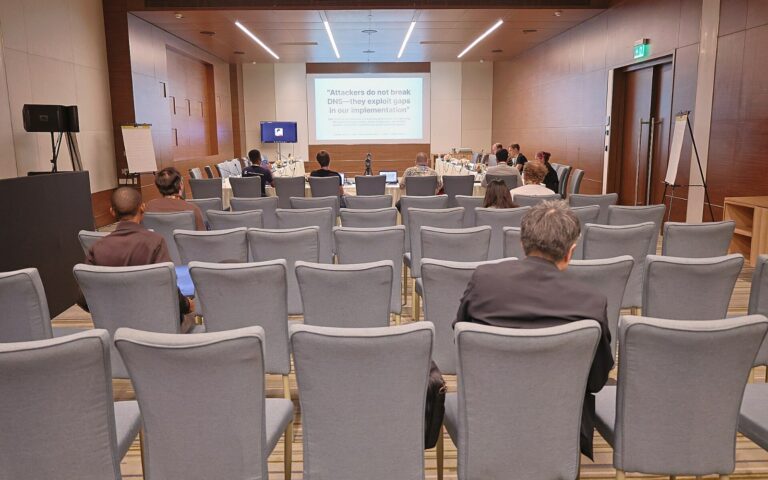 Salle de conférence vue du fond, rangées de fauteuils gris en partie occupées, orateurs à une table sous un écran de projection