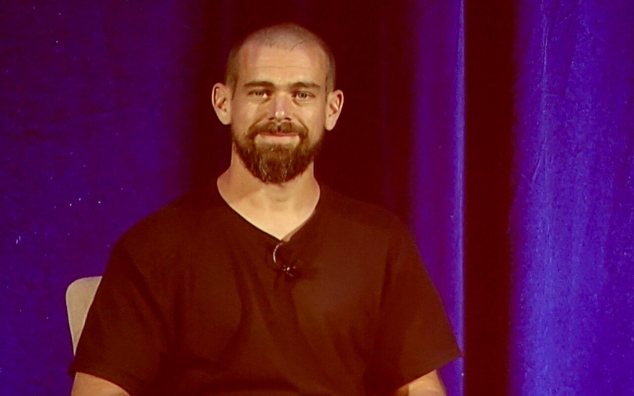 Jack Dorsey, barbu et souriant, assis sur scène devant un rideau violet lors d'une conférence en 2018