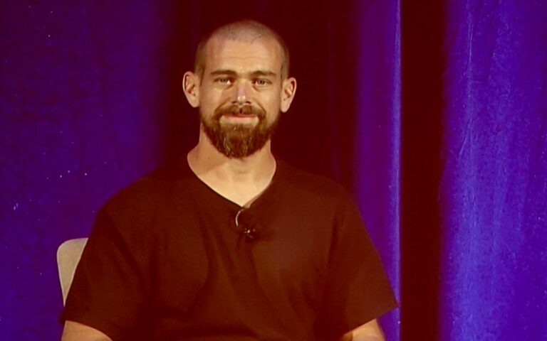 Jack Dorsey, barbu et souriant, assis sur scène devant un rideau violet lors d'une conférence en 2018