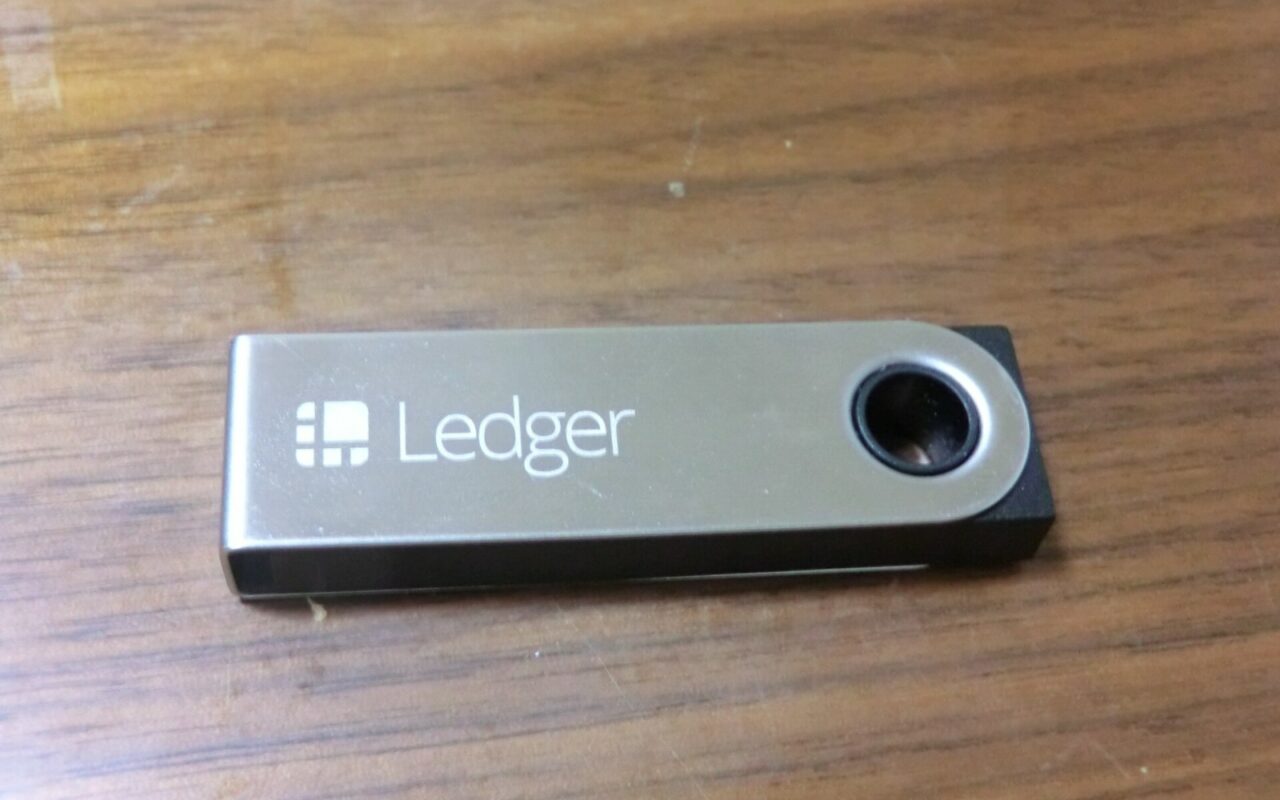 Un portefeuille matériel Ledger Nano S fermé, en métal brossé gravé du logo Ledger, posé sur une table en bois.