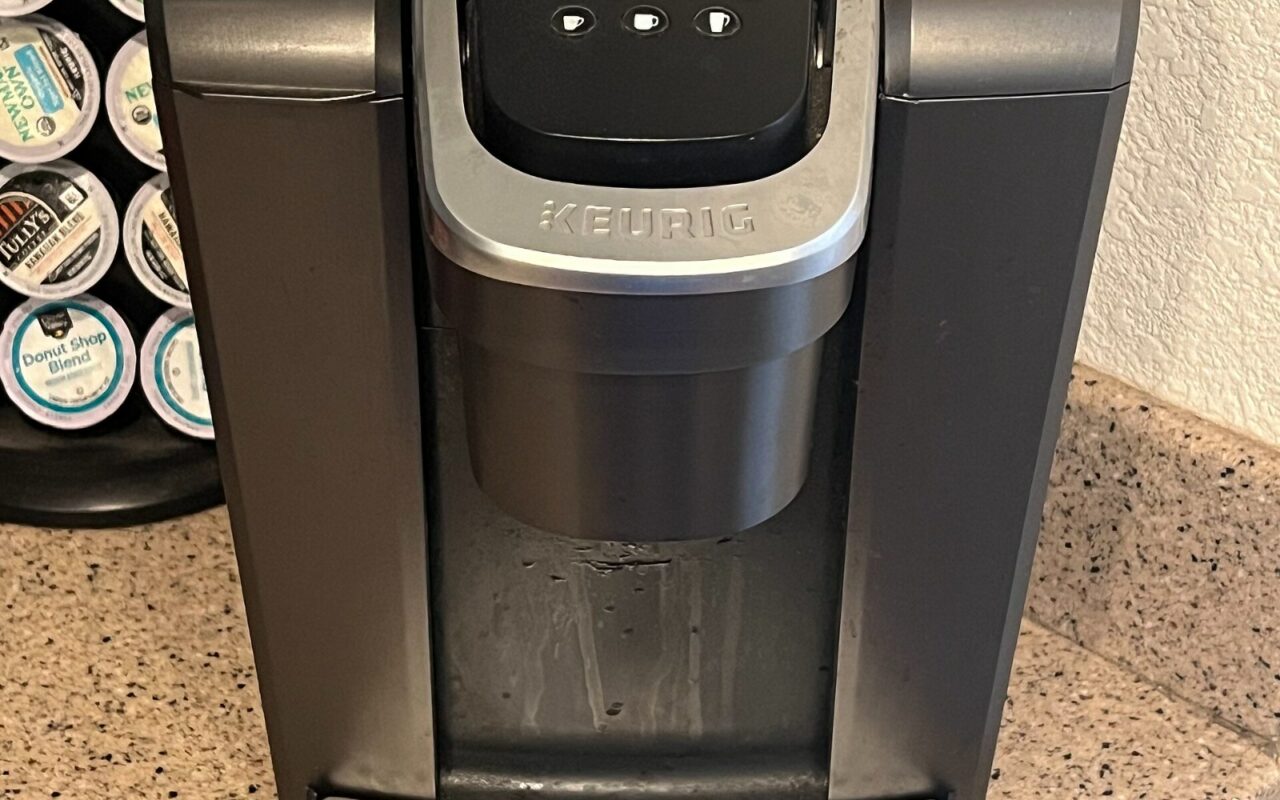Gros plan sur une machine à café à capsules Keurig grise et noire posée sur un plan de travail, à côté d'un présentoir de capsules.