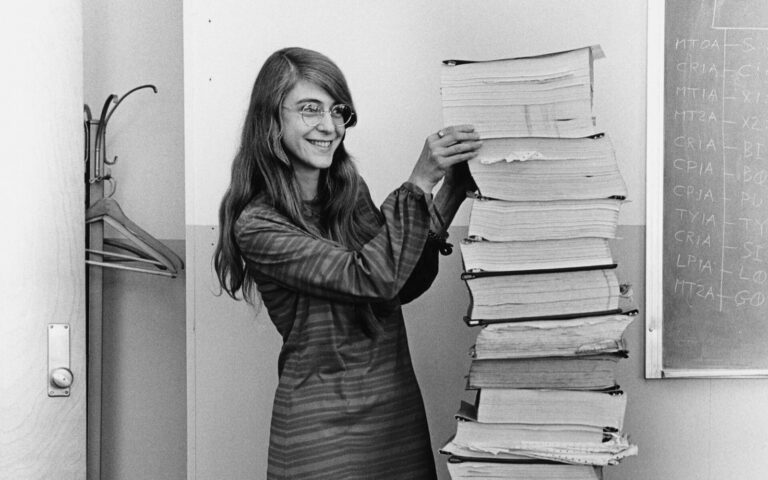 Margaret Hamilton en 1969, souriante, debout à côté d'une pile de listings du logiciel d'Apollo aussi haute qu'elle