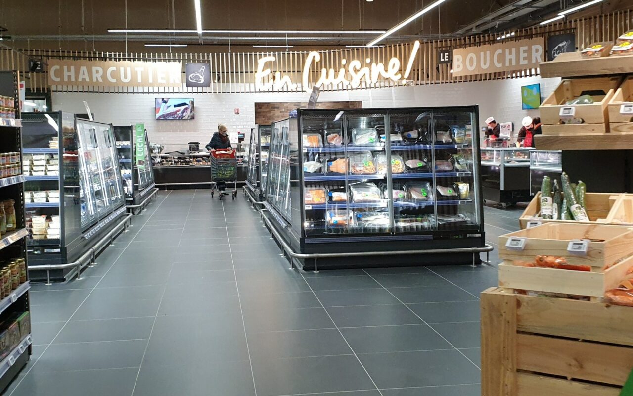 Allée d'un supermarché Intermarché bordée de meubles réfrigérés vitrés remplis de produits frais, sous les enseignes charcuterie et boucherie.