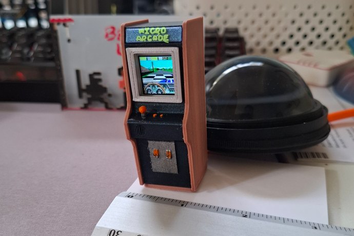 Borne d'arcade miniature orange et noire affichant un jeu en couleurs, posée près d'une règle graduée
