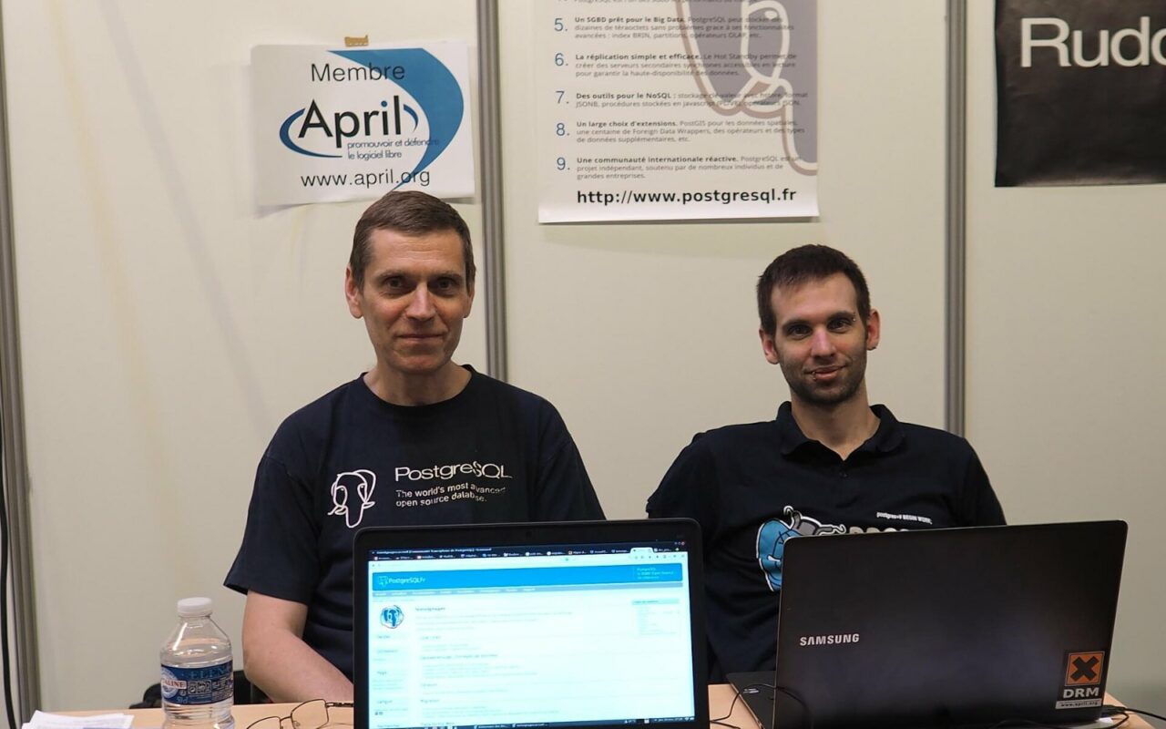 Deux bénévoles en tee-shirt PostgreSQL, assis derrière des ordinateurs portables sur un stand associatif d'un salon informatique.