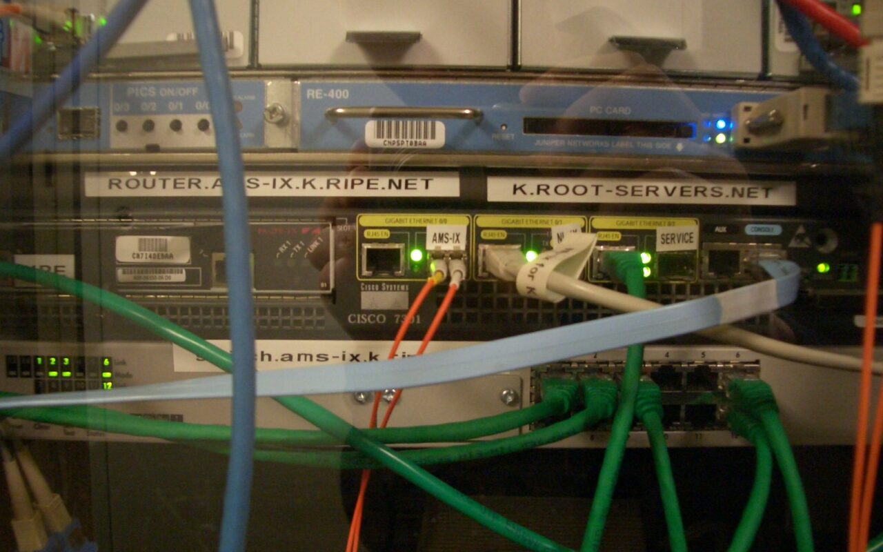 Baie informatique avec des équipements étiquetés k.root-servers.net et des câbles réseau verts et bleus