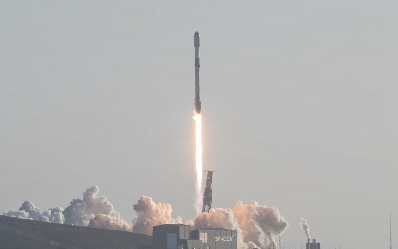 Une fusée Falcon 9 décolle dans un panache de fumée au-dessus d'un hangar marqué SpaceX, sur la côte californienne