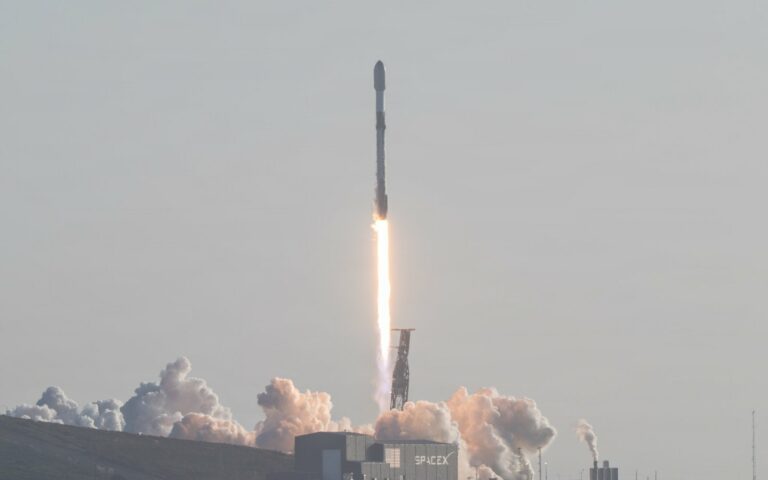 Une fusée Falcon 9 décolle dans un panache de fumée au-dessus d'un hangar marqué SpaceX, sur la côte californienne