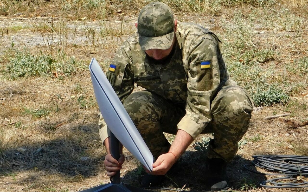 Un soldat ukrainien en tenue de camouflage installe une antenne Starlink sur un pied, dans l'herbe sèche