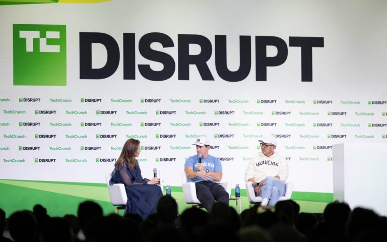 Trois intervenants assis sur la grande scène de TechCrunch Disrupt, sous un écran géant au logo Disrupt, devant le public