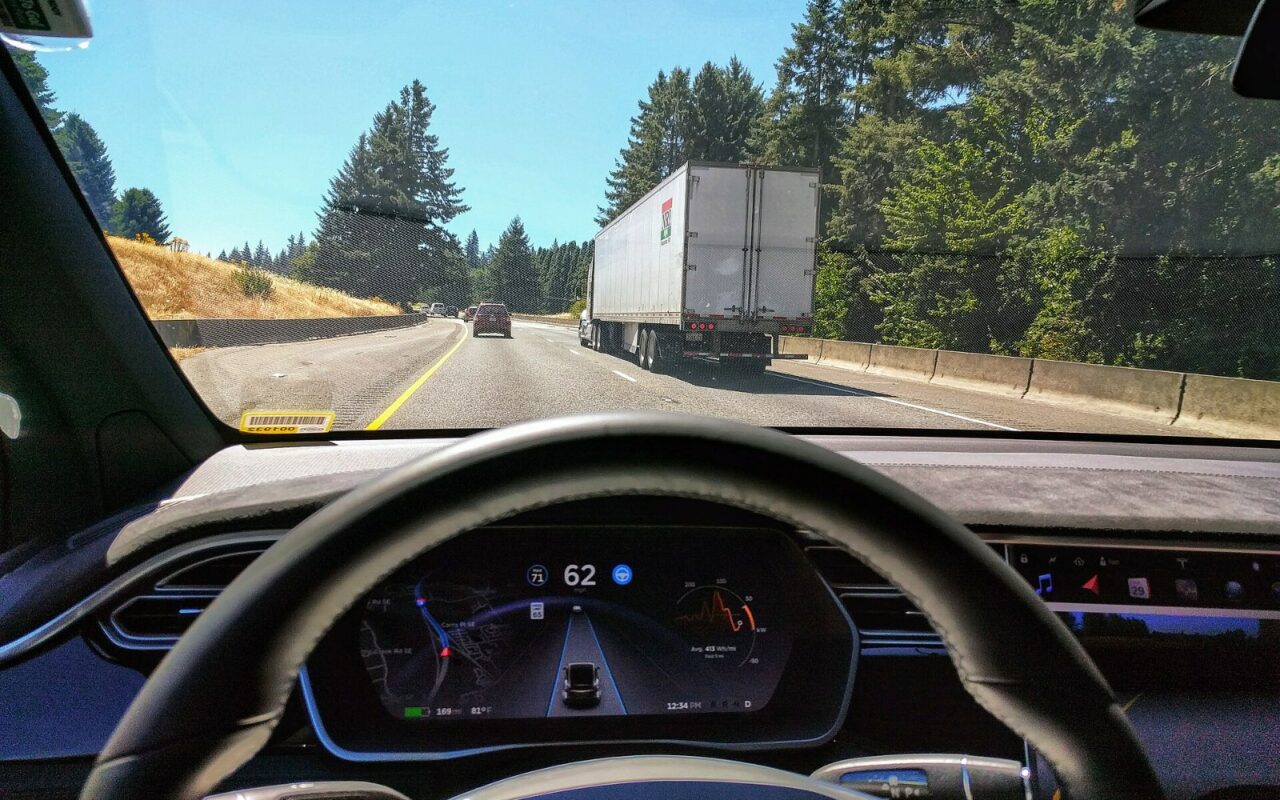 Vue depuis le siège conducteur d'une Tesla Model X sur autoroute : le volant, l'écran d'instruments et un camion sur la voie de droite.