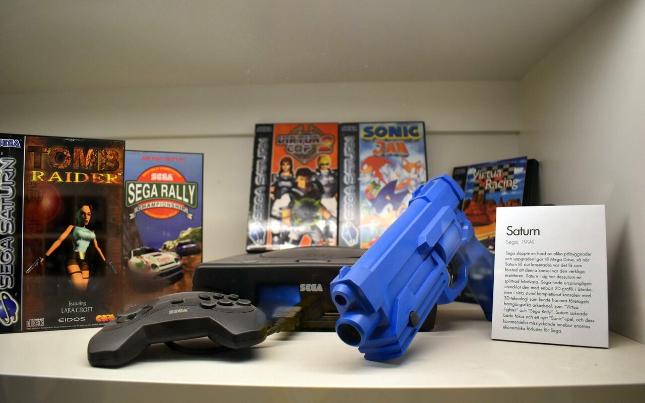 Vitrine de musée avec une console Sega Saturn, sa manette, un pistolet bleu et la boîte de Tomb Raider montrant Lara Croft